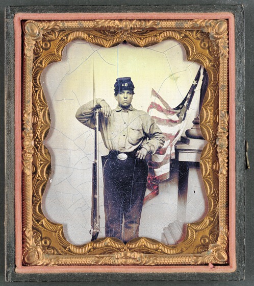 Unidentified soldier in Union uniform with bayoneted musket in front of painted backdrop showing American flag and column pedestal