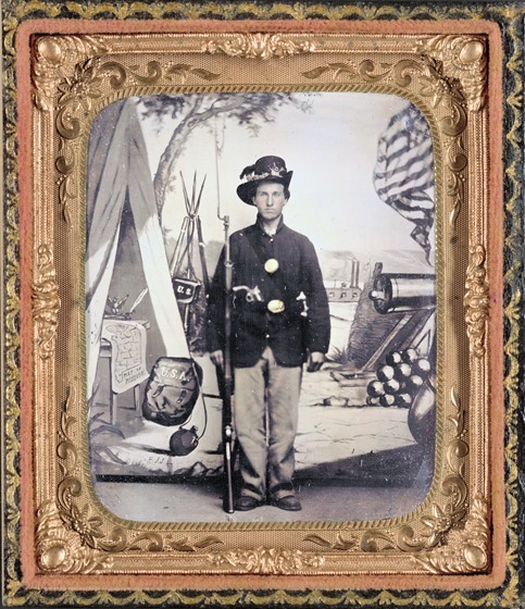 Unidentified soldier of 33rd Missouri Infantry Regiment with bayoneted musket and revolver in front of painted backdrop showing weapons and American flag at Benton Barracks, Saint