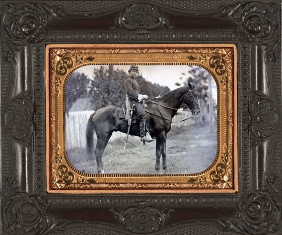 Unidentified soldier in Union first lieutenant's uniform and kepi with cavalry saber atop horse in front of fence, building, and woods in picture case