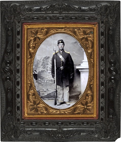 Unidentified soldier in Union frock coat and shoulder scales with bayoneted musket, cap box, and cartridge box in front of painted backdrop showing lake and trees in frame