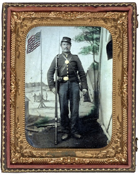 Unidentified soldier in Union uniform with bayoneted musket, canteen, cartridge and cap boxes in front of painted backdrop showing military camp with American flag in photo case