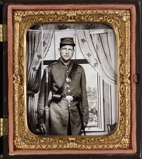 Unidentified soldier in Union uniform with bayoneted musket in front of painted backdrop showing a window with curtains in photo case