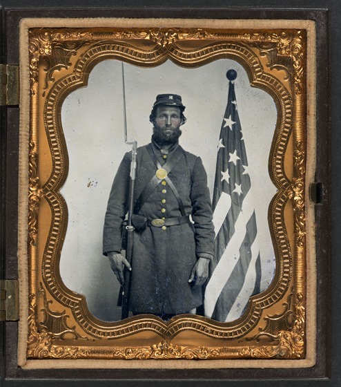 Unidentified soldier in Union uniform and Company H cap with bayoneted musket, cap box, and Volunteer Maine Militia (VMM) belt buckle in front of American flag in case